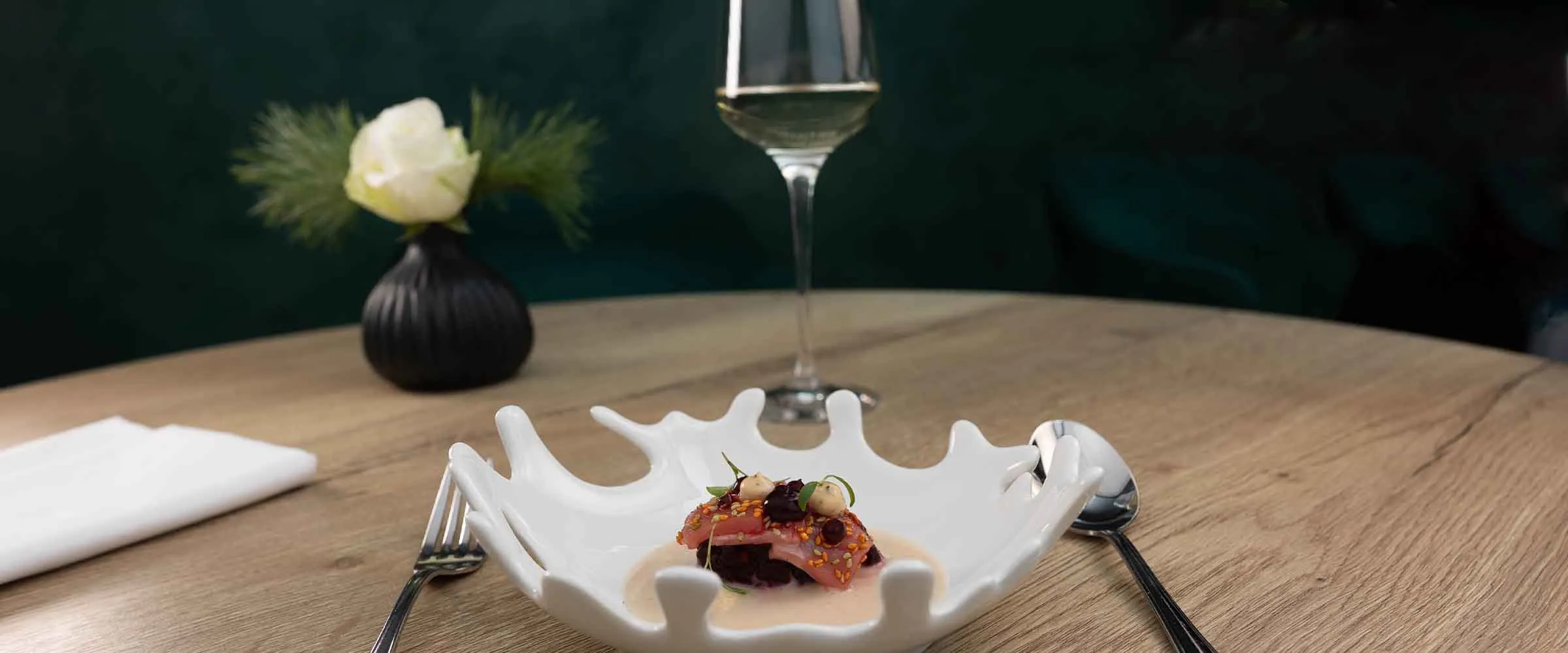 Fein angerichtetes Gericht auf weißer Porzellanschale, dahinter ein Glas Weißwein und eine weiße Rose in schwarzer Vase